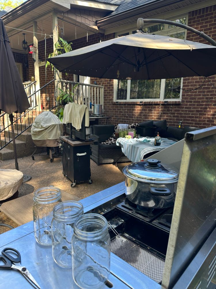 Grill with a pot of water, patio table in background with quarts of green beans and a table with a 24 quart pressure canner.  