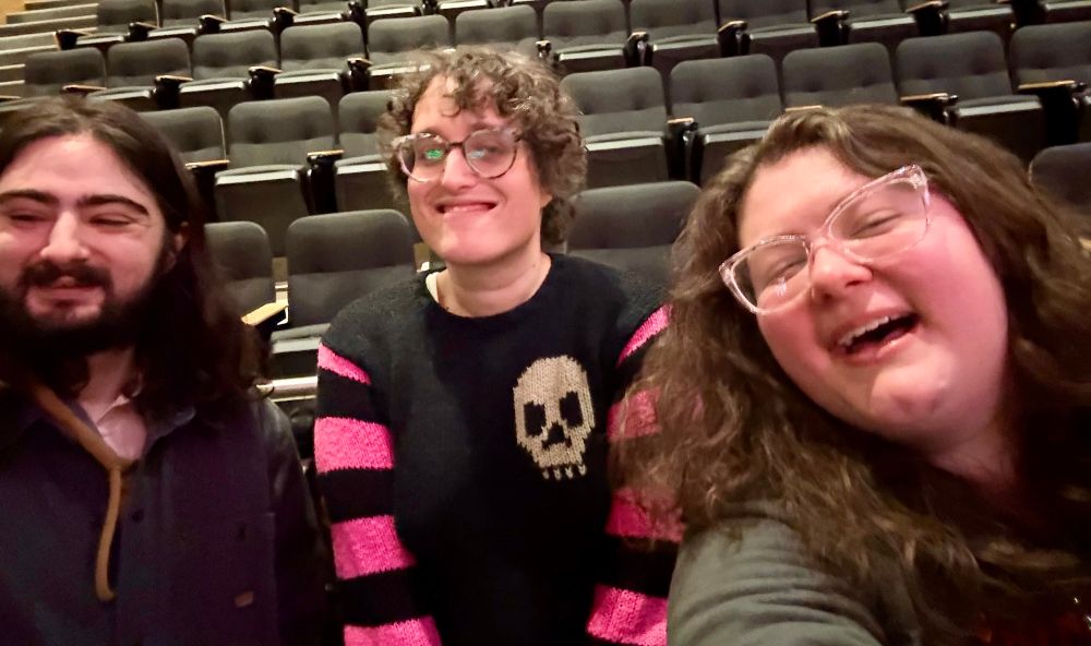 A selfie with my partner, director Jane Schoenbrun, and myself respectively from left to right. My partner has long straight black hair with facial hair (and a very cute smile). They're wearing a grey buttoned jacket over a brown hoodie. Schoenbrun stands between us, smiling. They are wearing a black long sleeve with pink stripes down both arms and a white skull on the left shoulder, as well as glasses. I'm on the right, smiling like an idiot. I have long wavy brown hair. I'm wearing clear pinkish glasses and a greenish grey long sleeve.