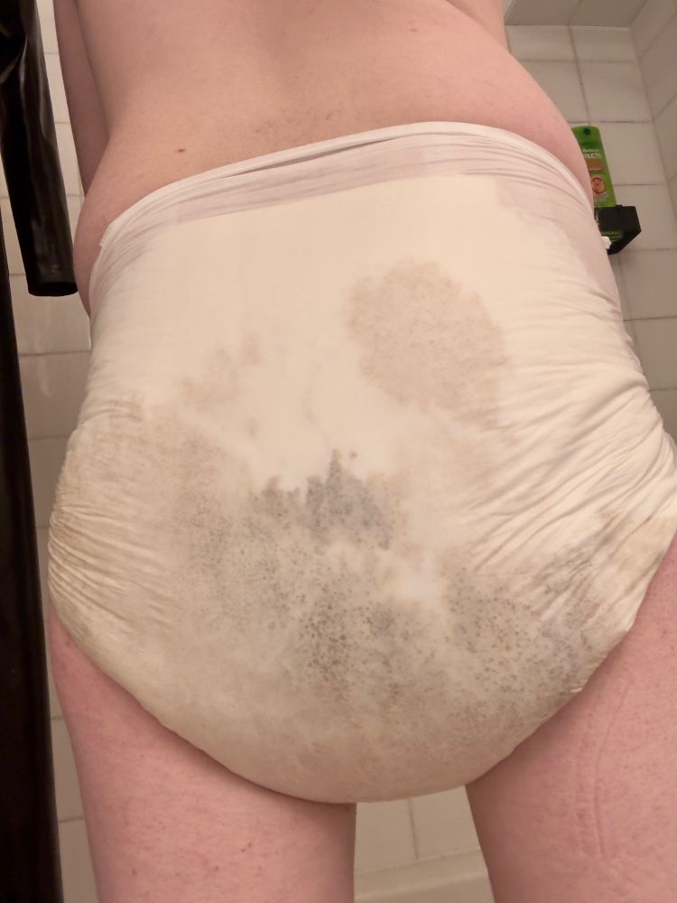 Rear view of someone wearing a white diaper. The bottom of the seat is heavy and full, dark brown stains covering it leaving nothing of its smell to the imagination 