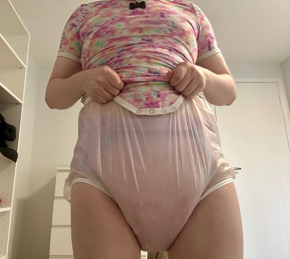 A close up of someone wearing a diaper. They have a pink tie dye onesie, plastic pants, and a lavender coloured trest. Even through the plastic pants you can see how stained and swollen the trest is, as it strains against the pants