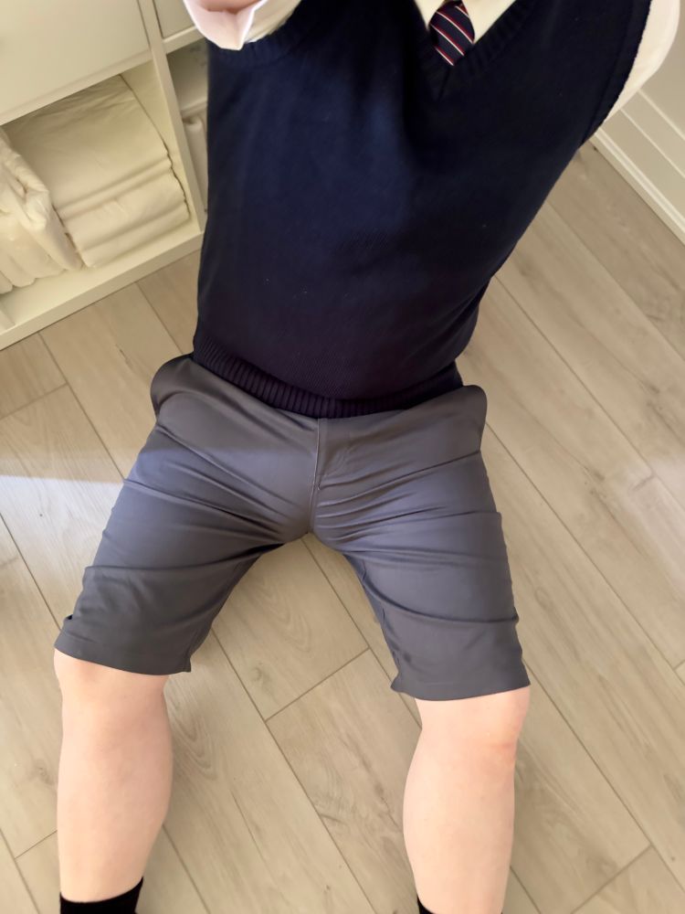 A person in a schoolboy uniform, sitting on the floor. They have tight grey uniform shorts, with an absurd diaper bugle pressing through very visibly. 
They wear a sweater vest and short sleeve white oxford shirt with a double Windsor tie.