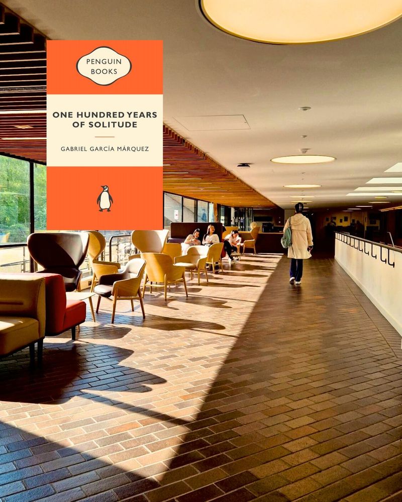 Photo of IOE building interior. A woman is walking towards the end of a corridor. There are chairs on the left hand side and squares of light are illuminating only the left side. Accompanying book is 100 Years of Solitude by Gabriel Garcia Marquez.