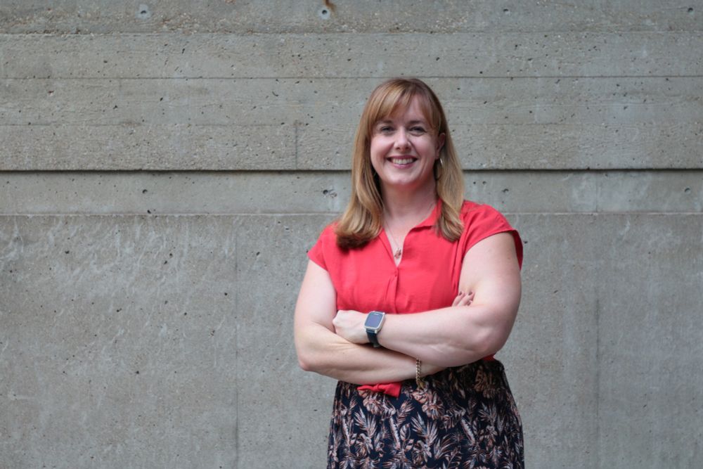 Sinead Harmey with crossed arms in a skirt and a red top in front of the IOE building's wall.
