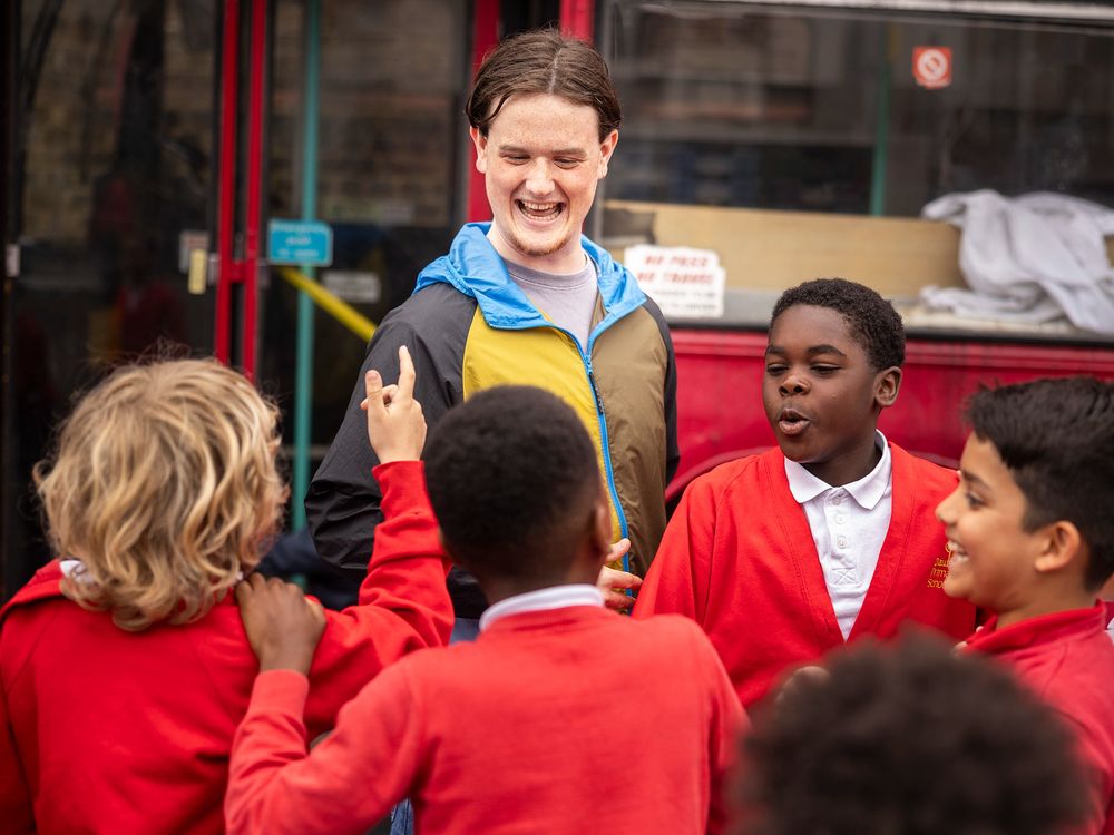 A young male teacher smiles with a group of boys in red school jumpers in the playground.