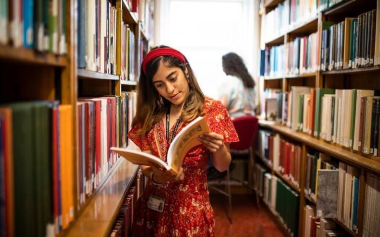 A woman holding a book and reading it, surrounded by shelves in the library.