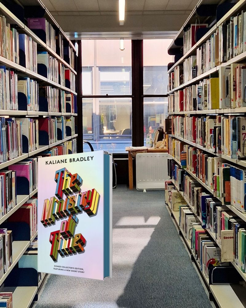 Photo of the IOE library between two shelves of books. The accompanying book is Ministry of Time by Kaliane Bradley. The book is silver with a colourful title.