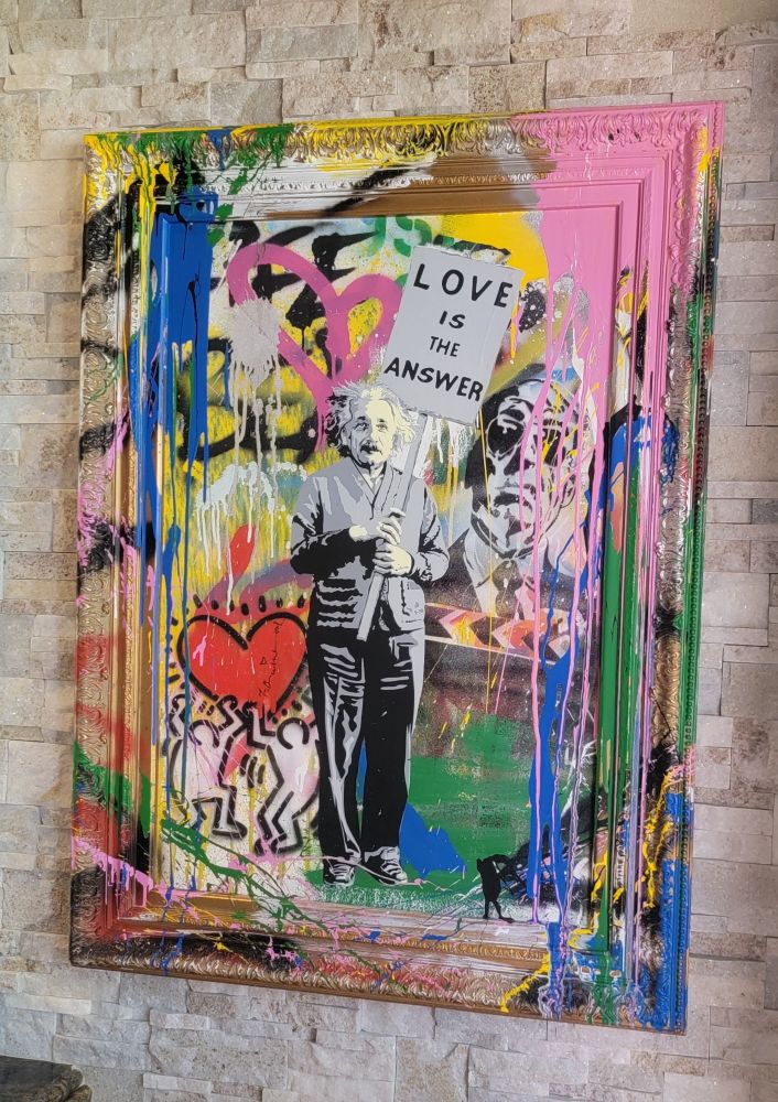 Bankey meets pop-art style artistic piece by Mr. Brainwash hangs on a stone wall. The work features an image of Einstein holding a "Love is the Answer" signs with cartoon figures holding up a large red heart below and beside him. Bold, splashed colors and outer edges of the canvas further make the image of Einstein and his sign stand out.
