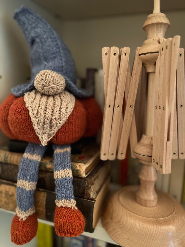 Una estantería, sentado sobre tres libros colocados horizontalmente hay un duende tejido a dos agujas. Tiene un sombrero puntiagudo algo doblado en color azul. Del ala del sombrero asoma una nariz redonda y gorda y una barba. El cuerpo es una gran calabaza naranja y, colgando de los libros, las piernas, colgando llevan medias a rayas azules y negros y zapatos naranja. 