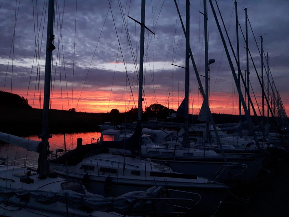Blick über Seggelboote in einem kleinen Hafen. Die Sonne ist schon fast untergegangen,  der Horizont ist leuchtend Orange.