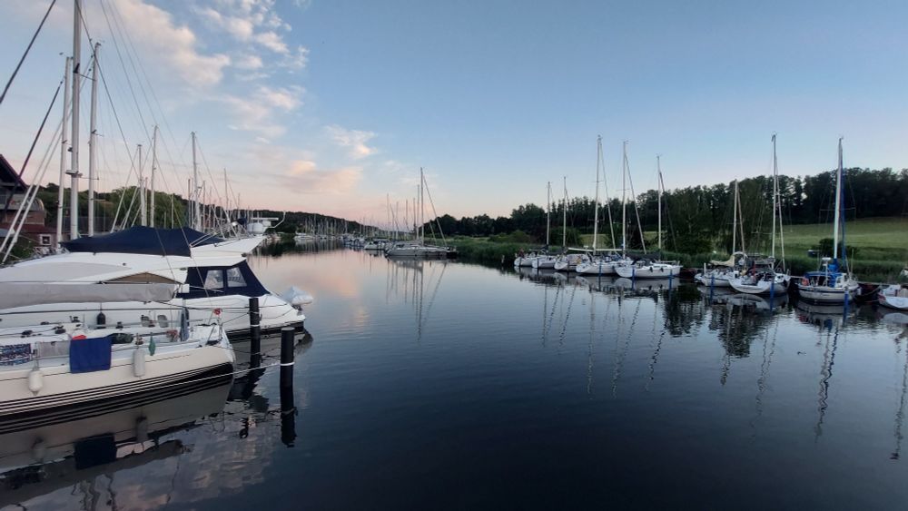 Man blickt von einer Brücke auf einen kleinen Naturhafen. Rechts und links liegen Segelboote und Sportboote.