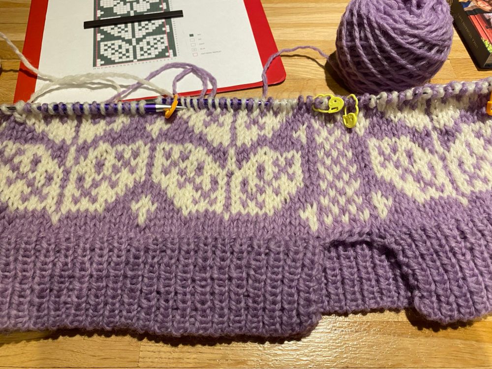 a knitting wip laying on a wooden table. it’s purple with white leaves, the second set of leaves is halfway finished. at the top are the chart and a ball of purple yarn. the pattern is the nordic bloom vest in rauma vams yarn