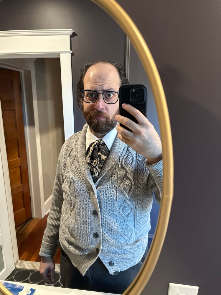 A middle aged man with a beard and longer hair wearing an 18th century cravat and a sweater making a dubious face