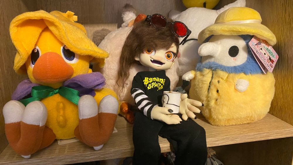 A picture of a customized Kabbit doll designed to look like the artist, wearing sweatpants and a Banana Splits t-shirt and holding a mug of tea while sitting among Plague Doctor and Final Fantasy plushies.