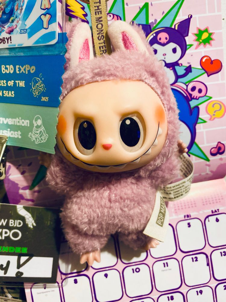 An accidentally aesthetic close-up of a purple Exciting Macaron Labubu hanging in front of a Sanrio calendar next to past convention badges.