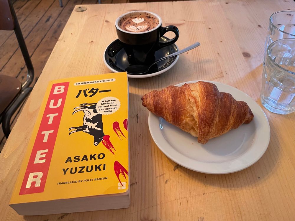 A wooden tabletop on which are placed a croissant, a cappuccino and a copy of Butter by Asako Yuzuki (translated by Polly Barton). The cover of Butter is yellow with a picture of a cow on it nose down,  smeared bloody finger prints on the right hand corner, the word butter in capitals in white on a red rectangle running top to bottom most of the length the left hand side, and Japanese script at the top that translates to butter.