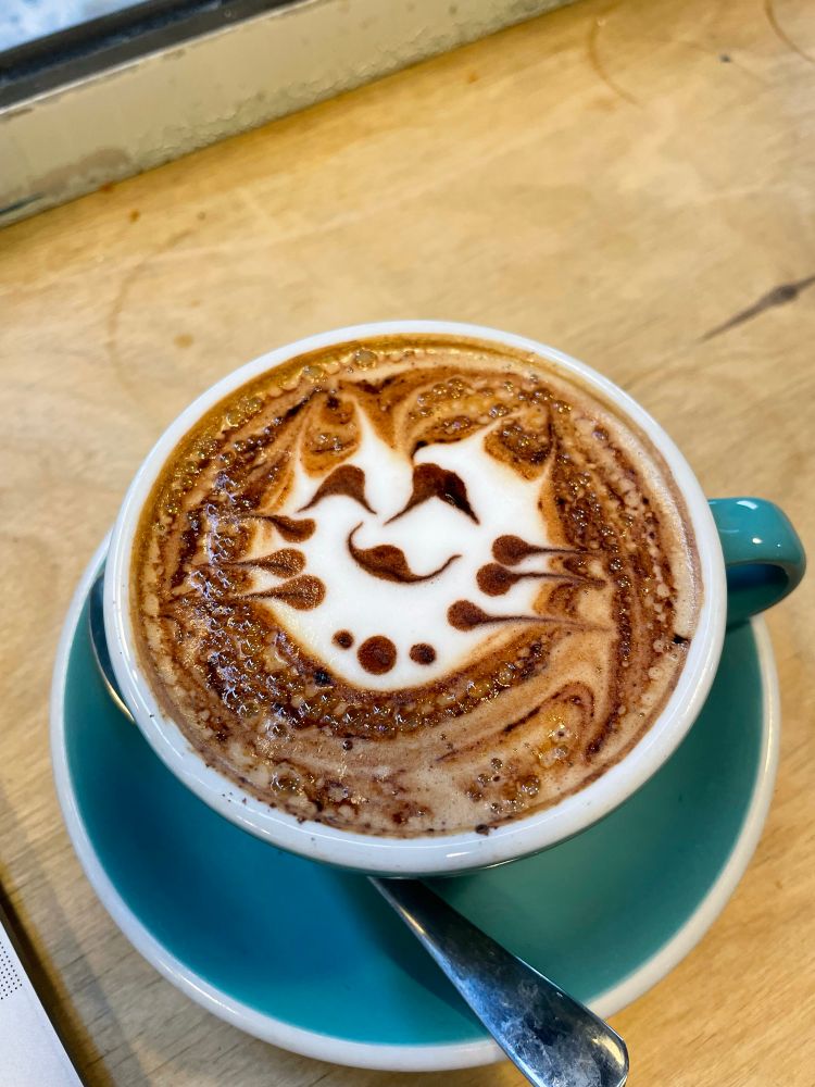 A coffee with a design that could be a cat on it but I’m not 100% sure. It’s resting next to a teaspoon on a turquoise saucer, which itself is on a wooden surface.
