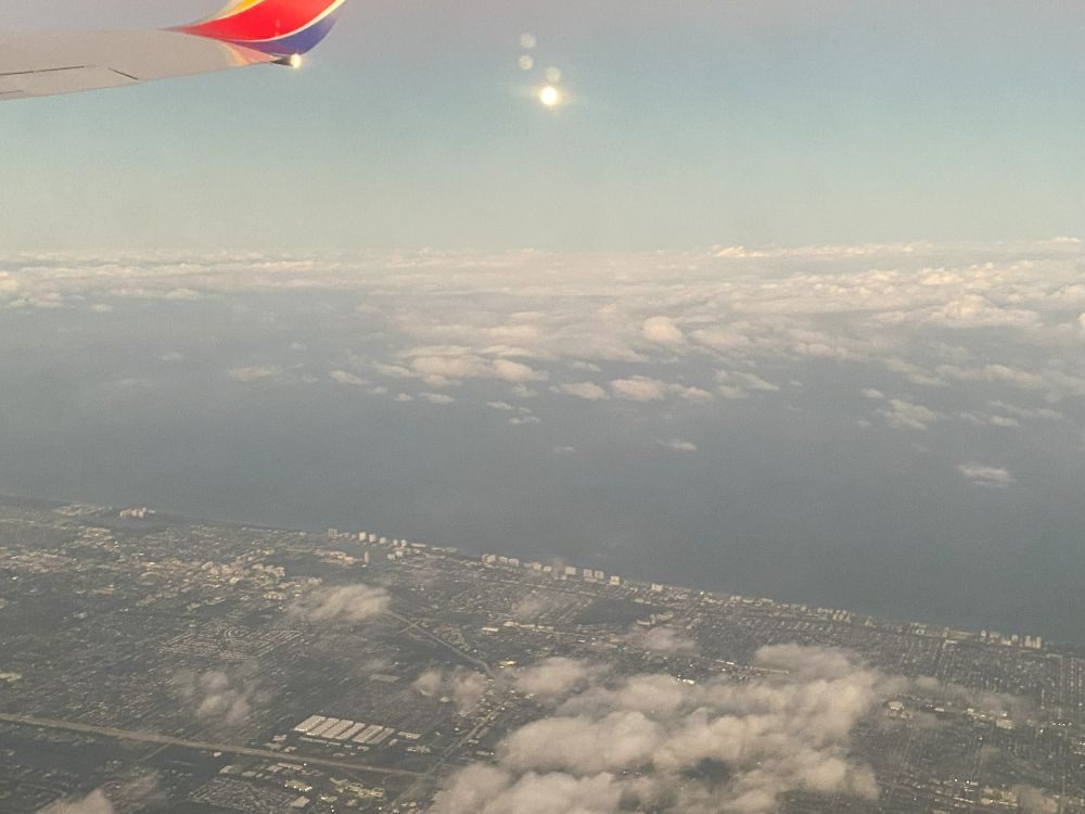 Looking out a plane's window down on the Florida coast, crammed full of highrises, buildings and roads. The coast butts up against the Atlantic Ocean, covered with patchy clouds. In the upper left corner is the tip of the plane's wing. In the center of the sky is the sun.