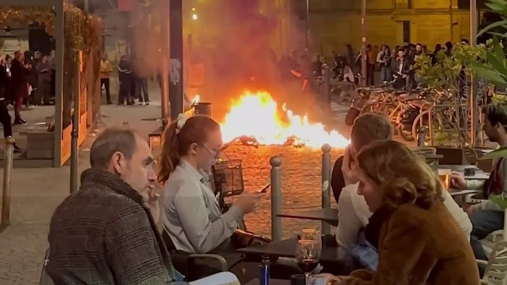 Image de manifestations en France. Au premier plan, des gens en terrasse. Au second plan, les manifestants avec un feu au centre dans la rue 