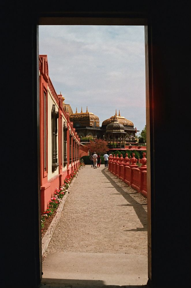 Looking through a doorway at a gravel walkway, with a red wall on one side and a red fence on the other. At the end stands the Palace of Gold with multiple gilded roofs.