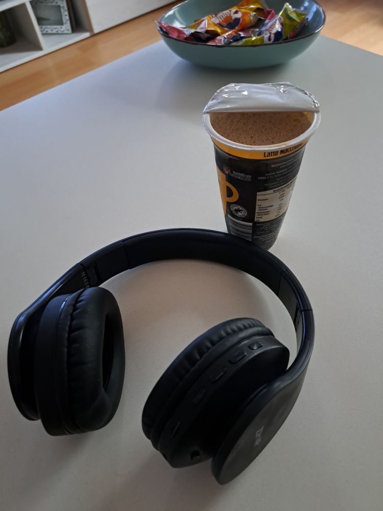 OverEar-Kopfhörer liegen auf dem Couch-Tisch... schräg dahinter steht ein geöffneter Becher Latte Macchiato.