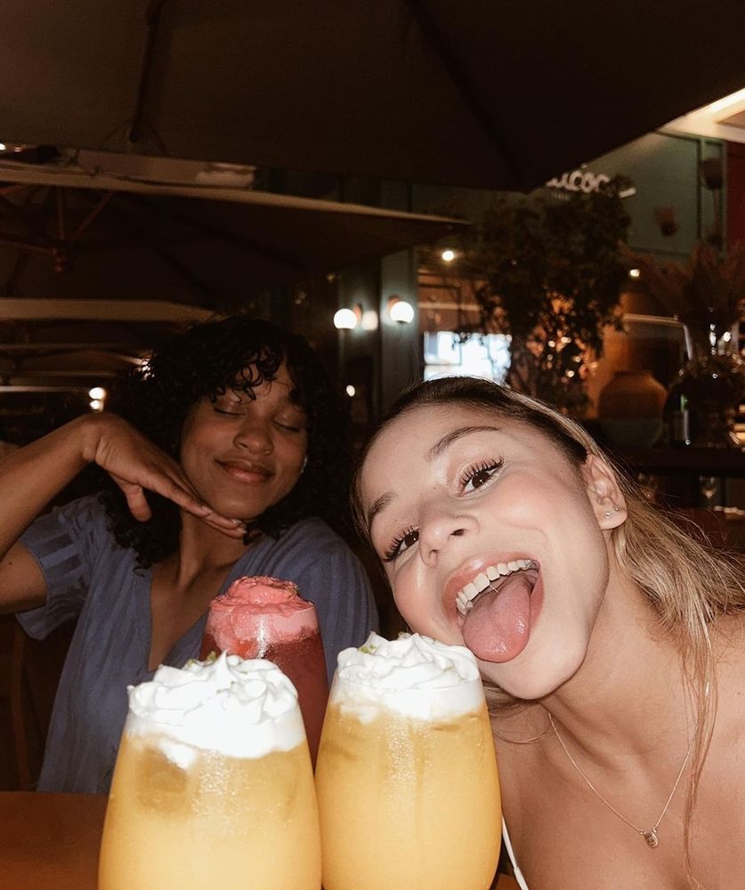 rebeca andrade de cabelo cacheado ao fundo e flavia saraiva fofa de com a língua para fora. Dois drinks amarelos na frente das duas. Aparentemente estão em um bar/restaurante.