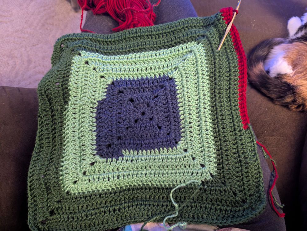 A square baby blanket in progress made of dark blue, light green, dark green, and a new row of red yarn.