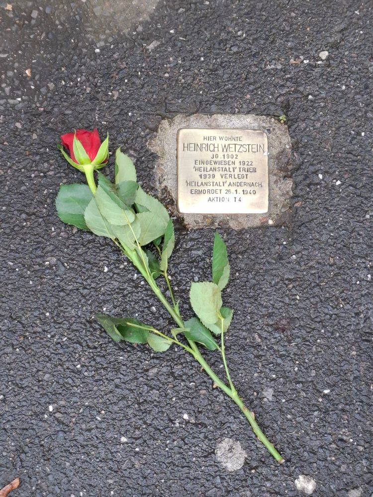 Rote Rose neben einem einzelnen Stolperstein mit folgender Aufschrift: "Hier wohnte Heinrich Wetzstein, Jahrgang 1902, eingewiesen 1922 'Heilanstalt' Trier, 1939 verlegt 'Heilanstalt' Andernach, ermordet 26.01.1940, Aktion T4"