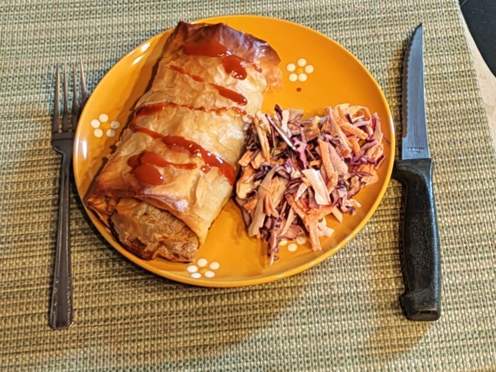 A large, golden phyllo pastry topped with a squiggle of hot sauce, beside a dollop of coleslaw, all on a yellow plate. 