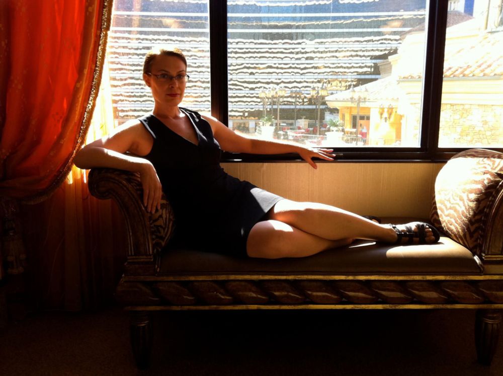 A woman lounging on an elegant sofa in front of a wide window with a red and gold drape. She is wearing glasses, and a tight, sleeveless chalk stripe dress in navy; her hair is up and her feet are scandalously up on the sofa cushion. 