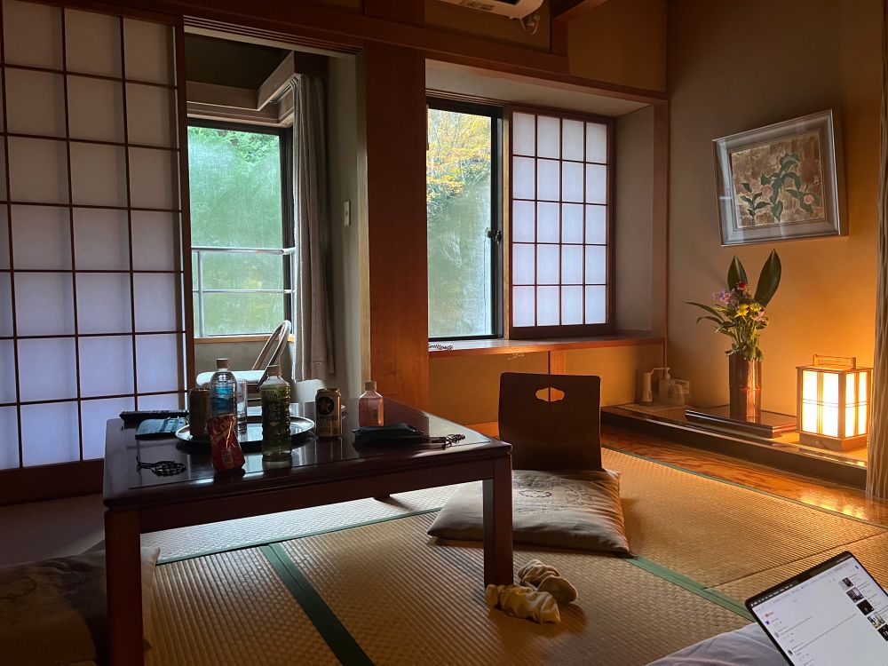 Slightly messy view of our room at Teiensaryo inn, we relaxed hard after a long trip! Bottles of various drinks are on the table in a traditional japanese room with paper screen doors and tatami mat floors. The light of the morning sun competes with a nearby lamp light to illuminate the dim room