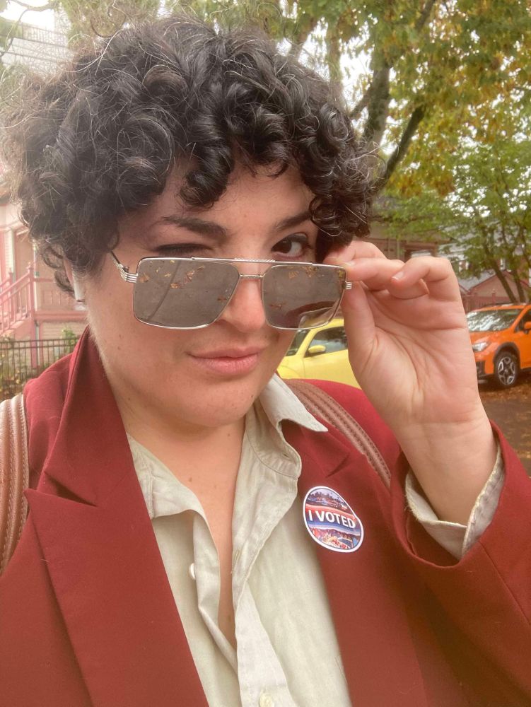 Molly as Agent Blanc winks at the camera over her sunglasses. She is wearing an "I Voted" sticker