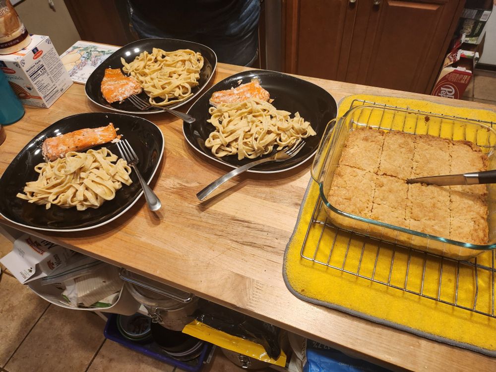 A kitchen island with three plates. Each plate has a piece of salmon and a pile of fresh pasta. To the right of the plates is an 8x8in glass baking dish with sliced biscuits