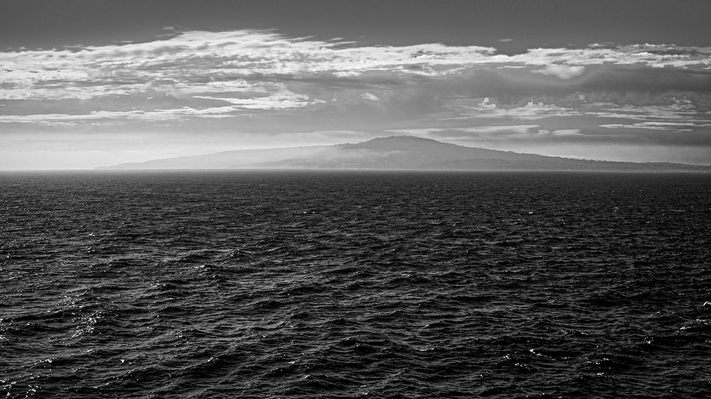 This is a black and white landscape photo I took at sea, facing a misty morning island in the Caribbean. The dark ocean catches light before the island, half-covered in curls of white sea mist. Above silver clouds stretch like frayed ribbons over a charcoal sky.