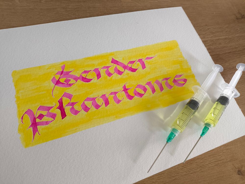 Photo of calligraphy reading Gender Phantoms. The text is bright pink on a yellow background, and two syringes are on top of the page