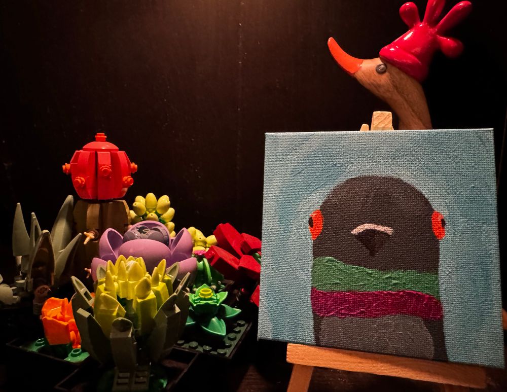 A cartoon pigeon with iridescent neck fluff on a light blue background set a top a small easel next to some Lego flowers and a wooden duck