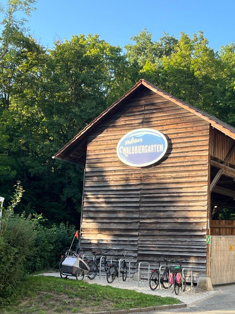 Großes Schild „Sudhaus Waldbiergarten“ auf Holzscheune im Wald, davor Fahrradabstellplätze mit Rädern verschiedener Art.