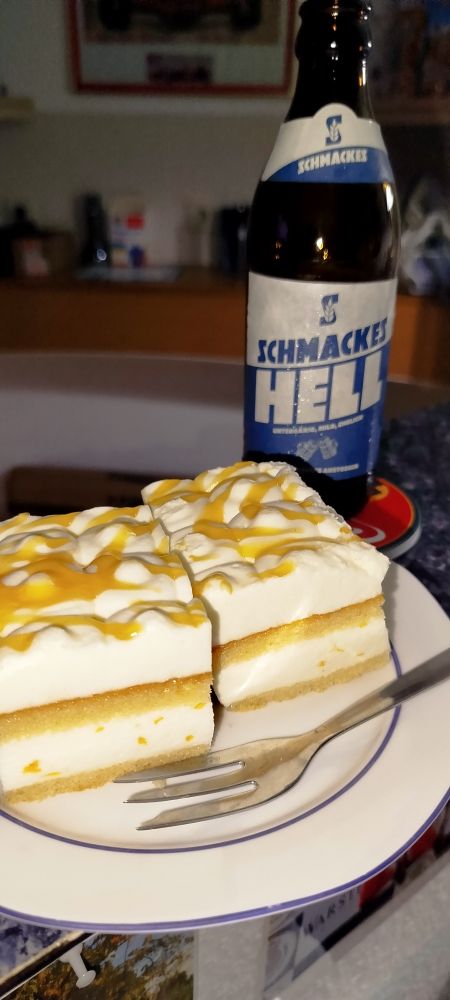 Ein Teller mit zwei Stück Käsesahnekuchen und dahinter eine Flasche Bier, Schmackes Hell. 