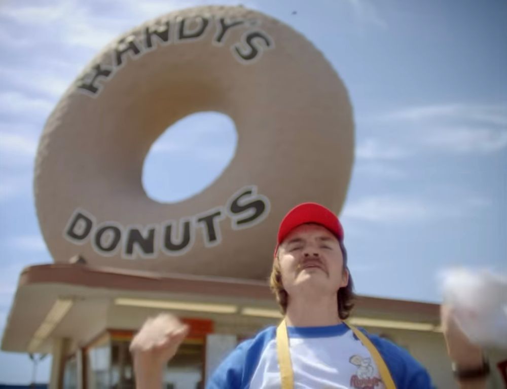 Still image from a Justin Timberlake , a man in a red hat and yellow apron is dancing in front of a giant donut that says “RANDY’S DONUTS” on it.