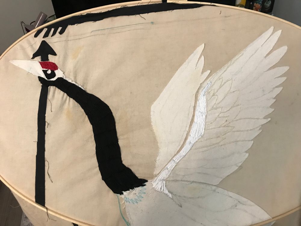 work in progress photo for a tapestry of a crane in a quilting hoop. It is a red-crowned crane, and has its neck, eye area, red crown and wings partially embroidered. Black arrows make up the "frame" of the tapestry.