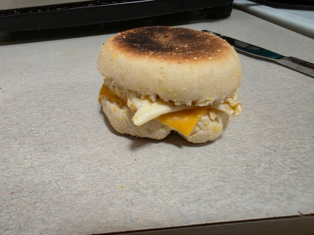 English muffin used to make a egg and cheese breakfast sandwich onto of a counter. The top of the English muffin is a little dark, but not yet in the burned category.