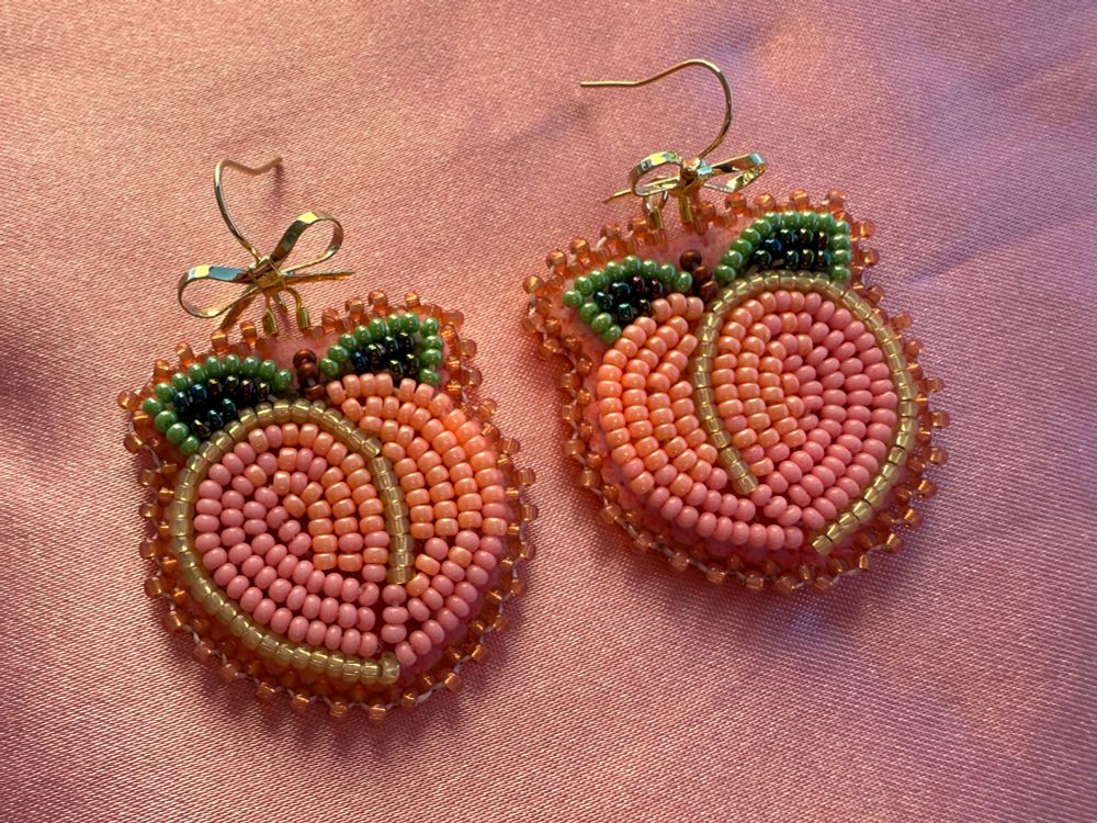 Flat stitch beaded earrings in a peach design. Gold plated hooks bent into shape of a bow. Peach and pink beads with green beads for the leaves. Laying on pink satin. 