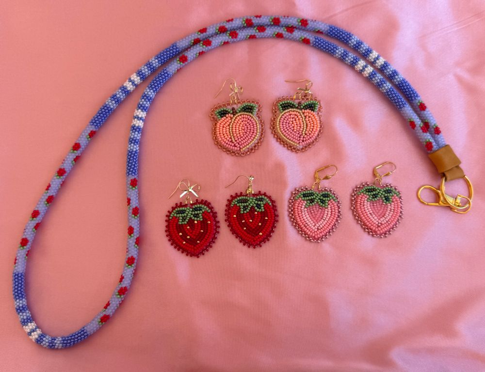 Blue Peyote stitched lanyard a strawberry design. One pair of beaded pink strawberry earrings. One red beaded strawberry earrings. Laying on pink satin. 