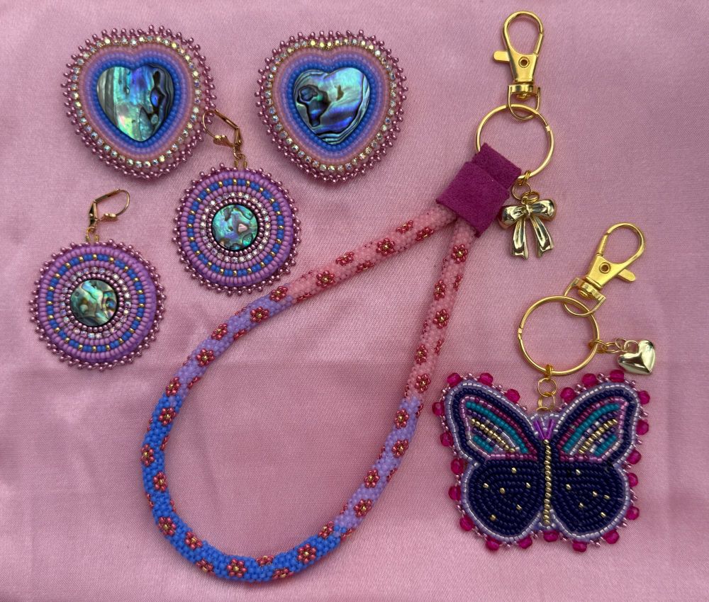 Beaded earrings with abalone centers, peyote stitched keychain, beaded butterfly. All made with pink, blue, and purple colored beads. Gold plated hooks and hardware. Beadwork is laying on pink satin. 