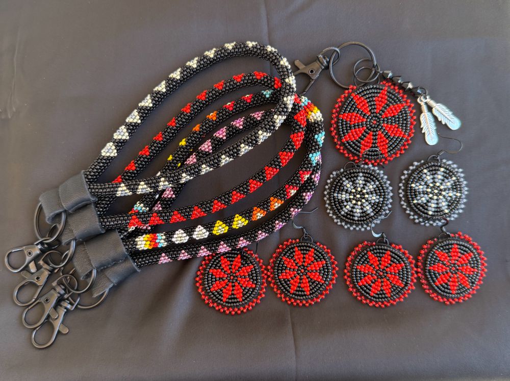 Set of Native beadwork in primarily black beads. Four peyote stitched loop keychains, Two pairs of black and red pendant earrings, one pair of black and grey pendant earrings, and one black and red pendant key chain. All laying on black satin.