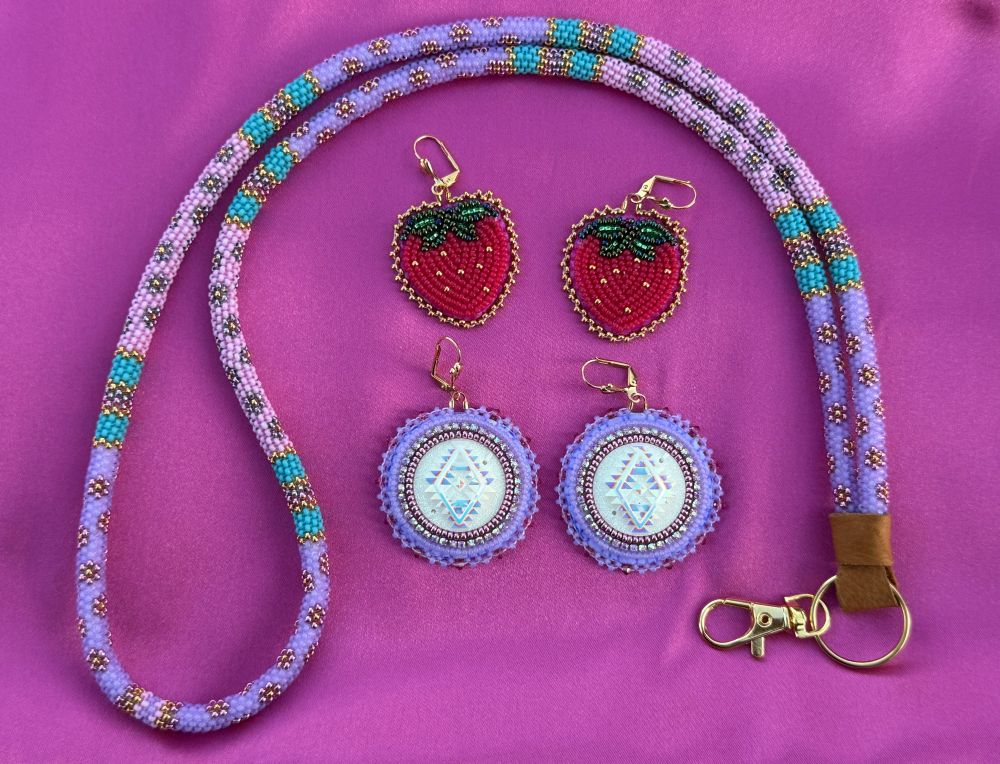 Native American beadwork. One purple and lilac beaded lanyard in a floral and striped peyote stitched design. One pair of light red beaded strawberry earrings. One pair of purple circular earrings with white southwest design resin center. Laying on magenta satin. 