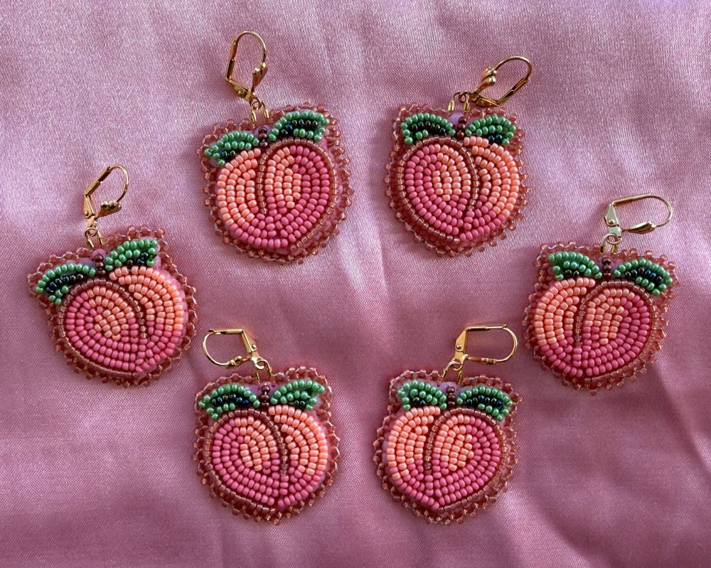Beaded peach earrings. Flat stitch design with pink and peach colors. Gold plated lever hooks. Laying on pink satin. 3 pairs laid out in a circle. 