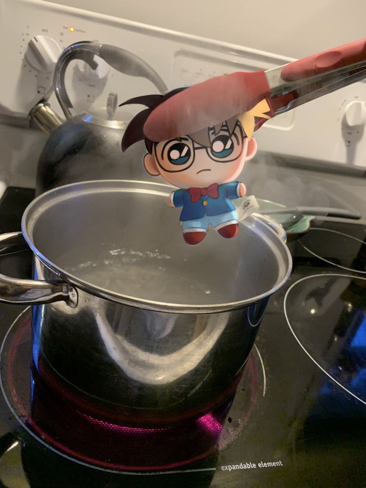 A plushie being held by a tong into a pot of boiling water on a stove. The plushie is drawn over to make it into a plushie of Edogawa Conan from Detective Conan.