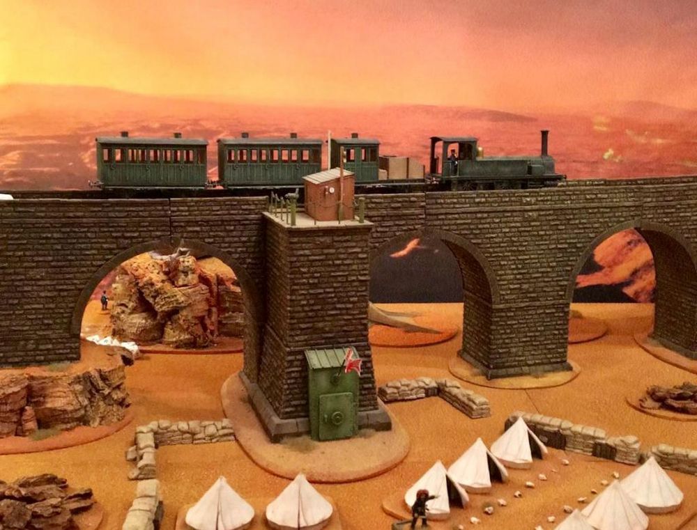 A green coloured train moves from left to right on a tall viaduct. There is a military camp below the viaduct surrounded by sandbags. 