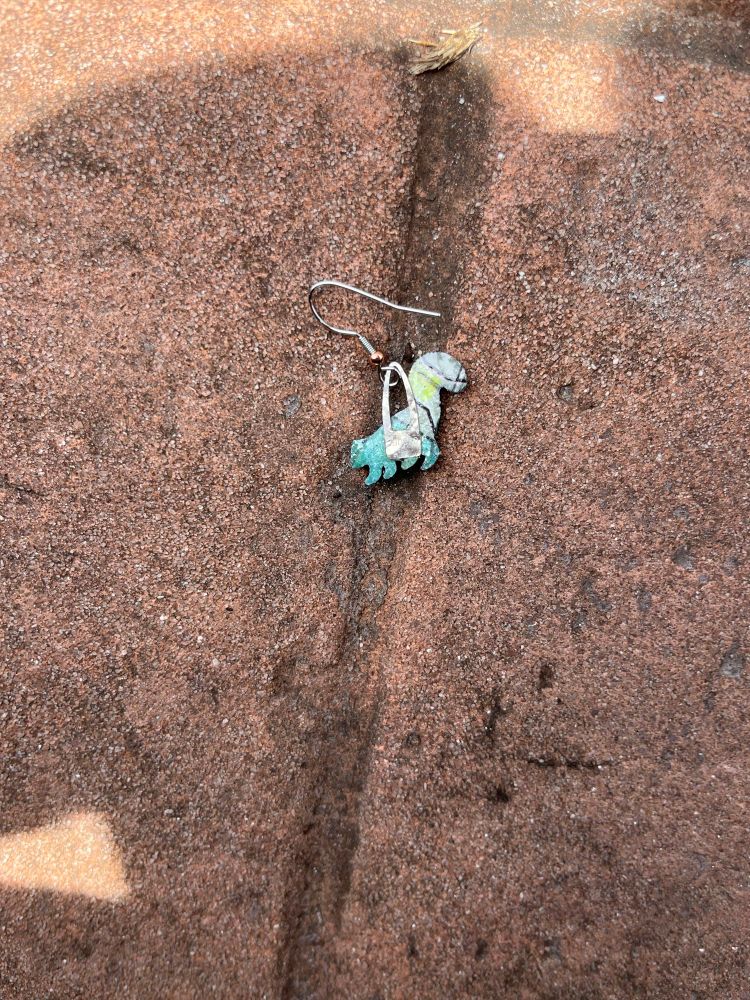 Close up: An earring (back side up) in the shape of a skunk with a wire loop laying on a brick driveway. 