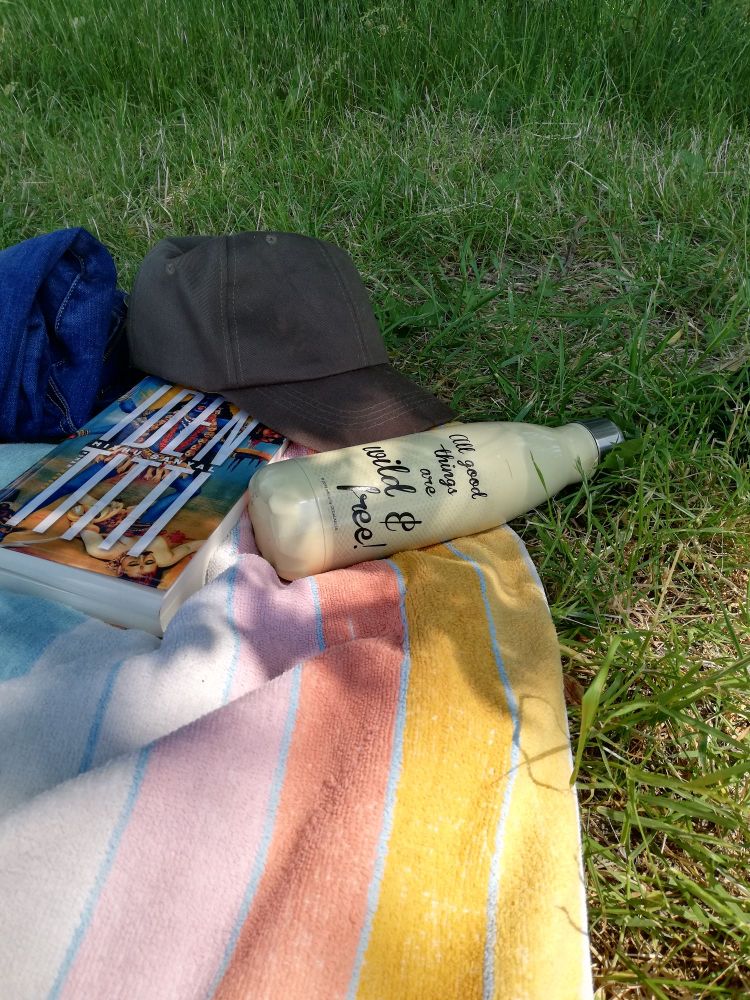 Ein gestreiftes Badetuch auf einer Wiese. Darauf ein Buch, eine Trinkflasche und eine grüne Schildkappe. 